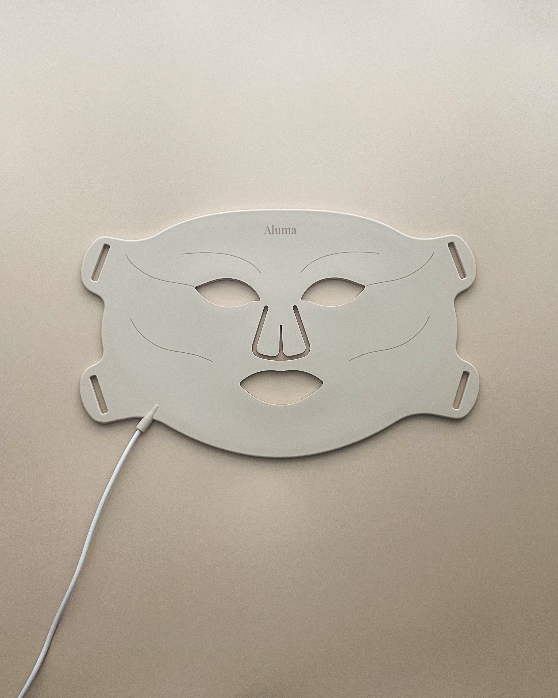 Aluma Aurora fleksibel LED-maske til ansigtet lagt fladt på beige baggrund med hvidt kabel, anti-age lysmaske til hjemmebrug.