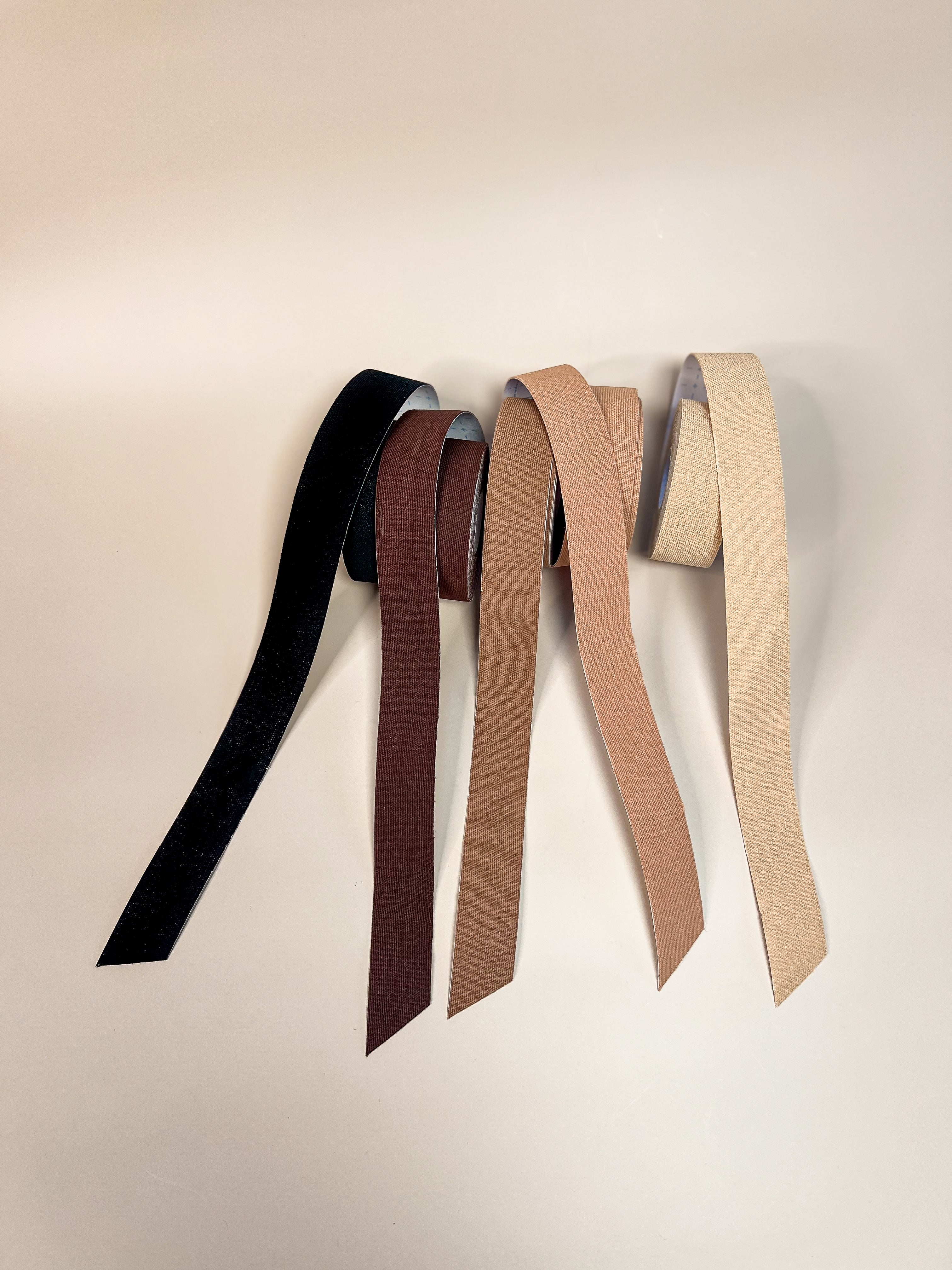 Four hanging strips of Aluma Contour face tape in black, deep brown and two beige tones, showing colour and cotton texture on a beige background.