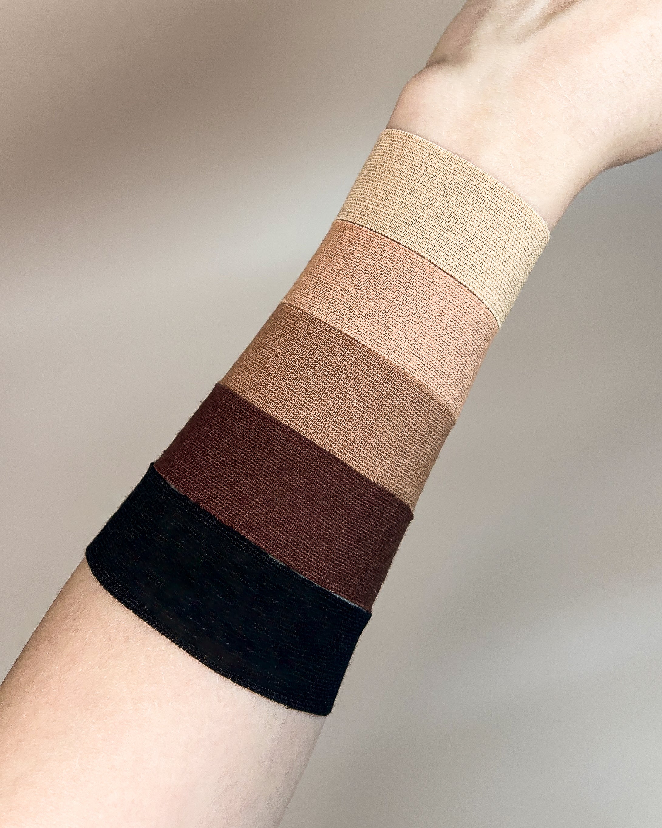 Four Aluma Contour face tape for sleep shades applied as wide bands on a forearm, from deep black to light beige, against a soft beige backdrop.