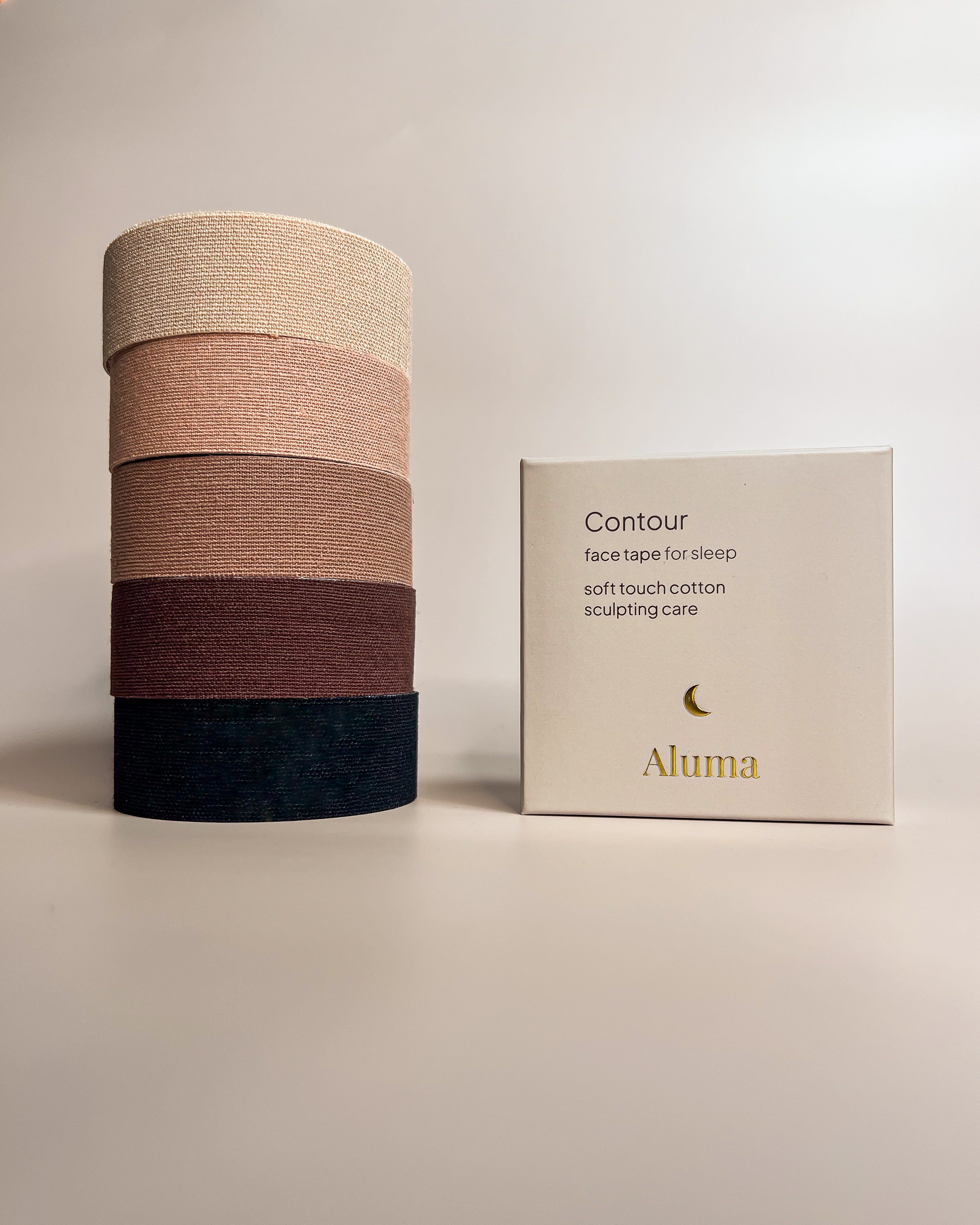 Four Aluma Contour face tape rolls stacked from light beige to deep black next to the Contour face tape for sleep box on a beige background.