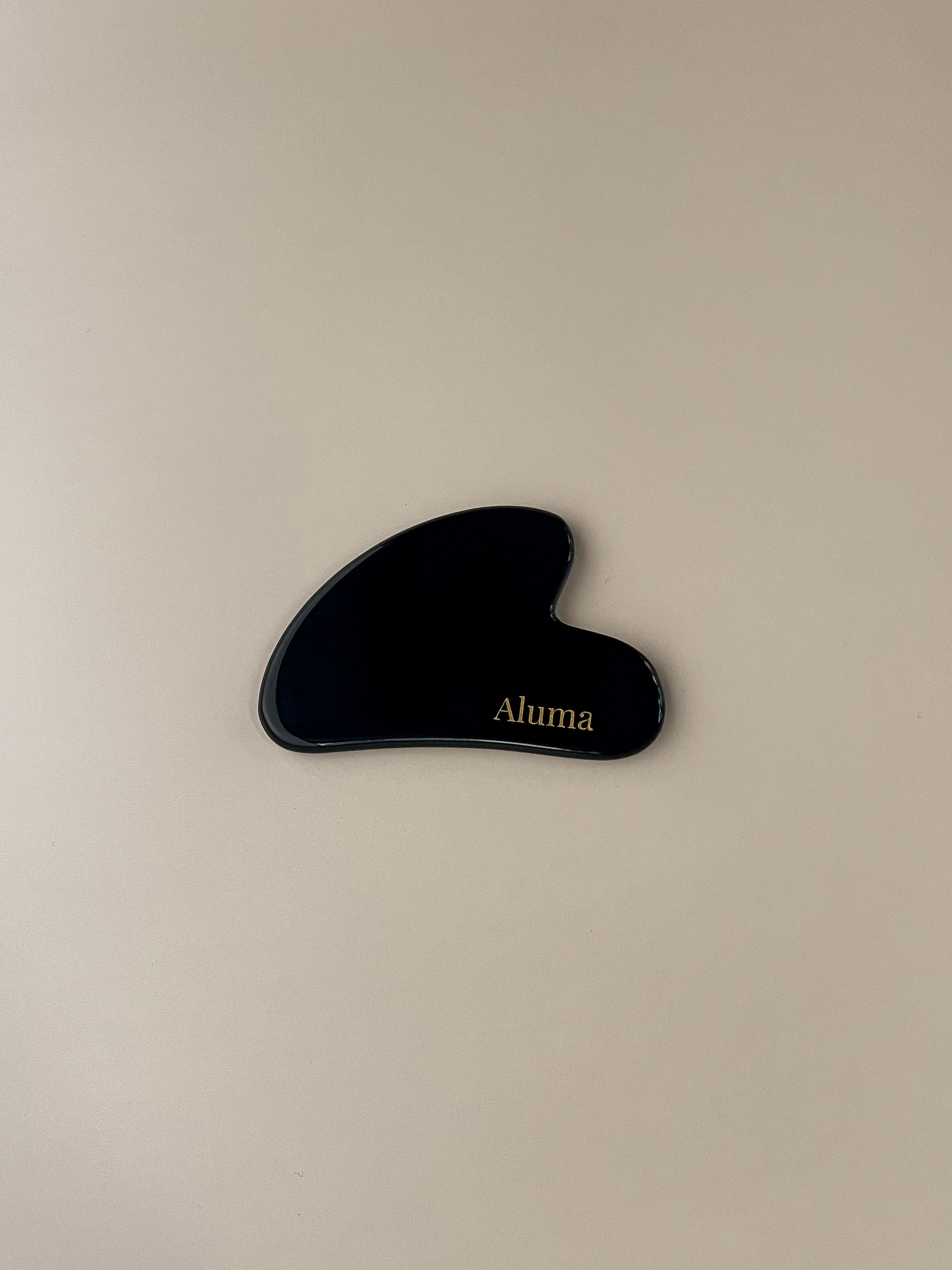 Yin gua sha i sort obsidian fra Aluma på beige baggrund, blank mørk sten der giver kontrast og dybde til ansigtsmassage og gua sha ritualer.