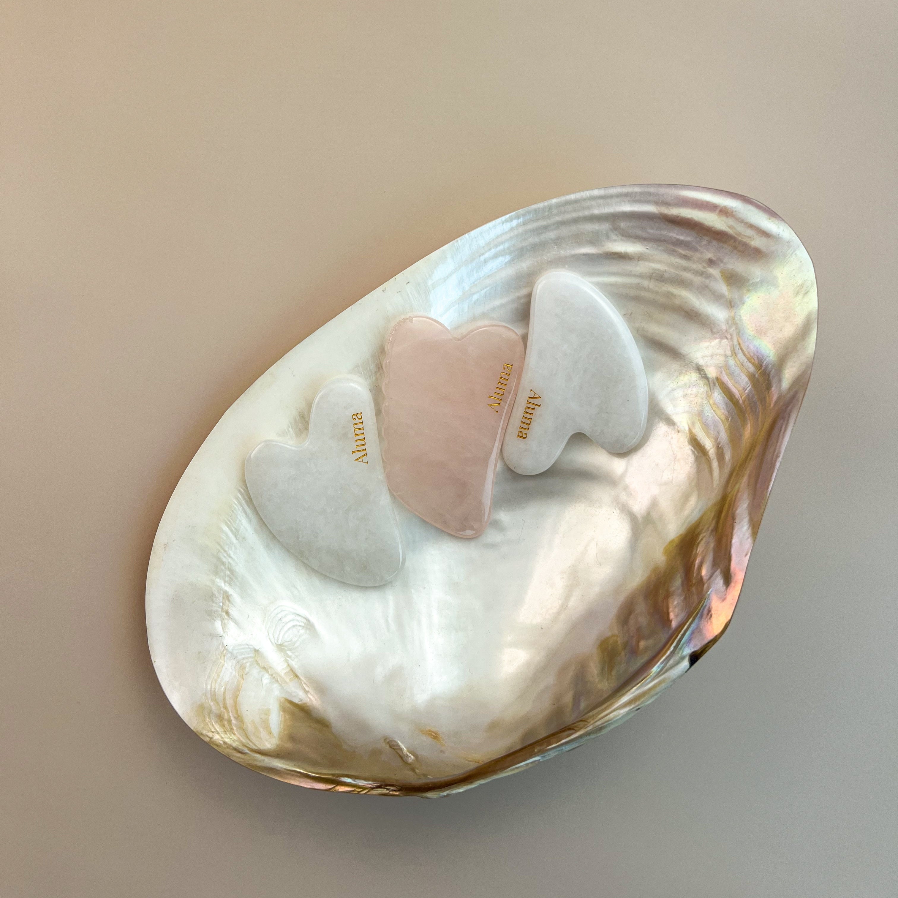 Tre Aluma Yin gua sha-sten i clear crystal, rosa kvarts og hvid jade placeret i en stor perlemorsmusling på beige baggrund.