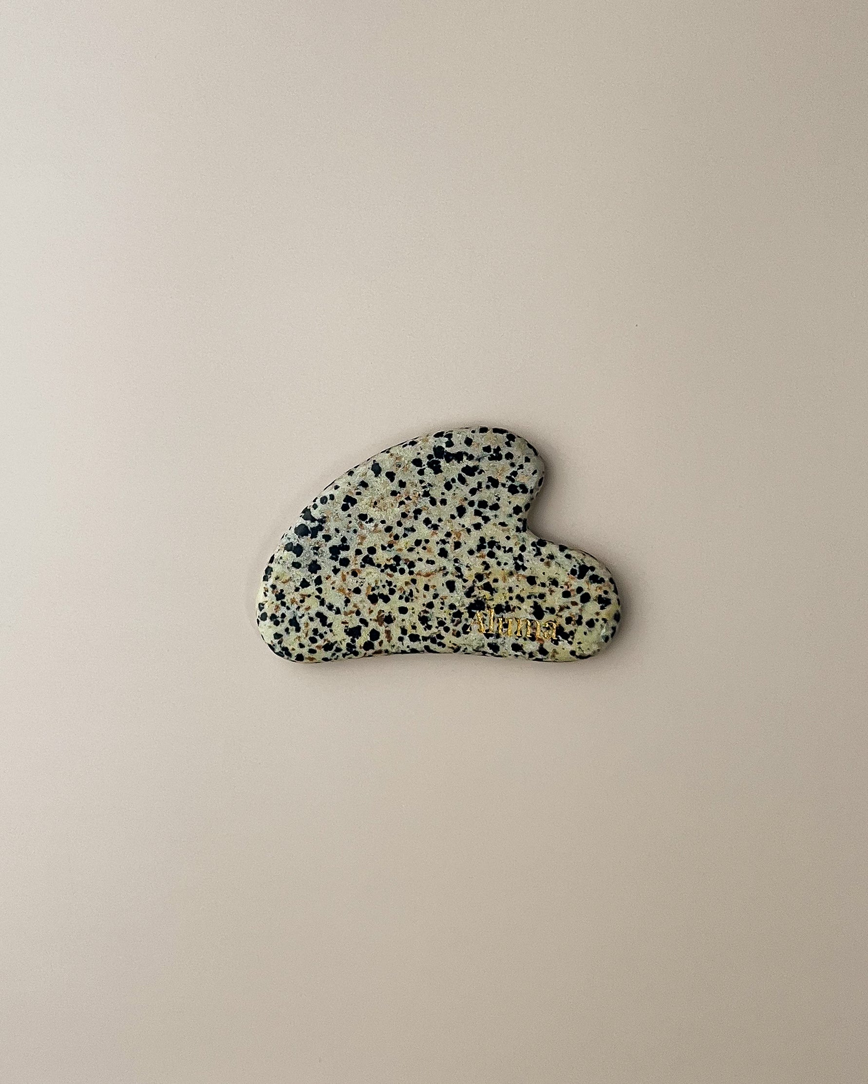 Yin gua sha i dalmatiner-sten fra Aluma på beige baggrund, lys sten med mørke pletter til blid ansigtsmassage og dekorative slow aging ritualer