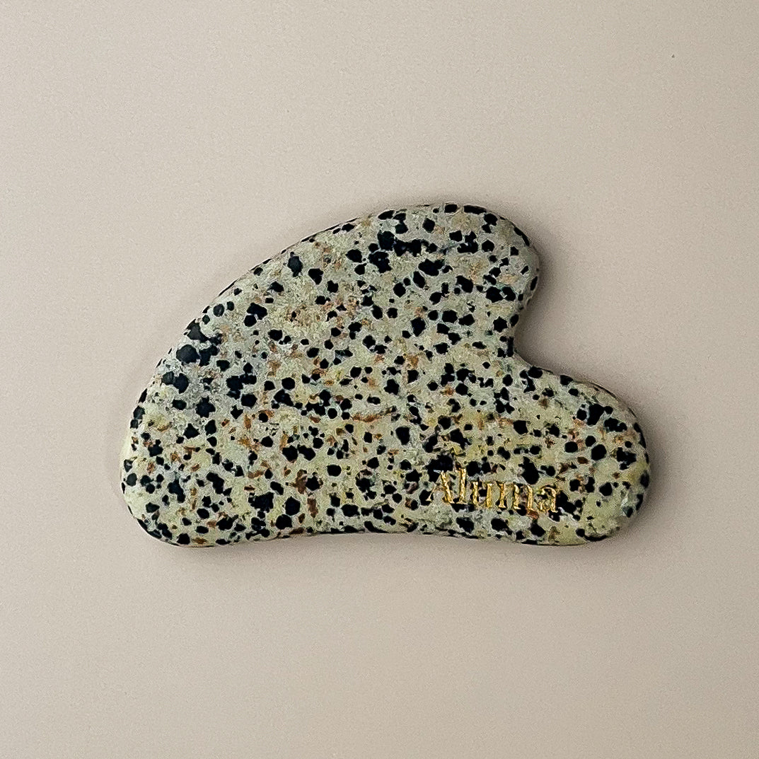 Yin gua sha i dalmatiner-jaspis fra Aluma på beige baggrund.