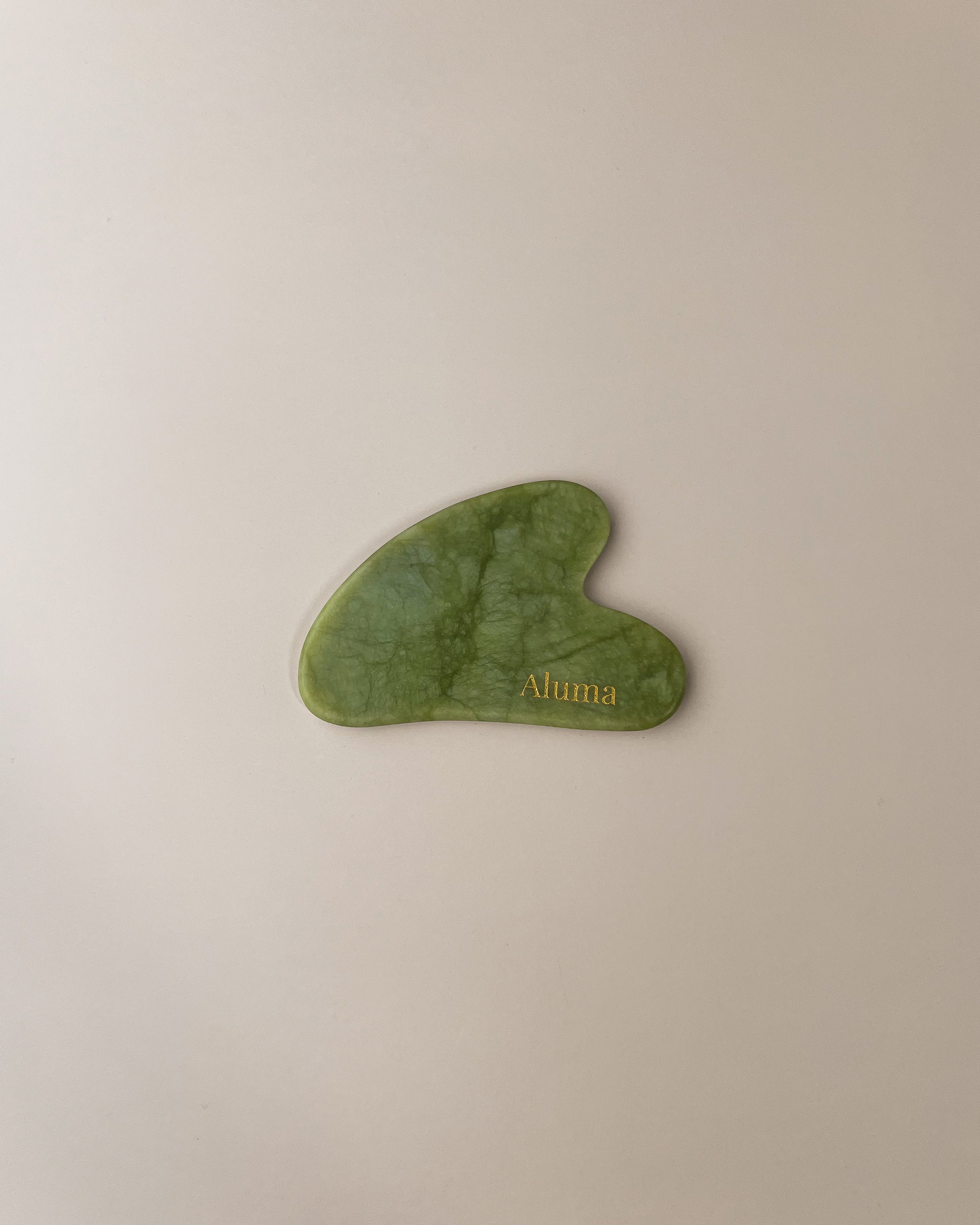 Yin gua sha i green nephrite jade fra Aluma på beige baggrund, klassisk gua sha sten til ansigtsmassage, spændingsløsning og opstrammende slow aging rutiner.
