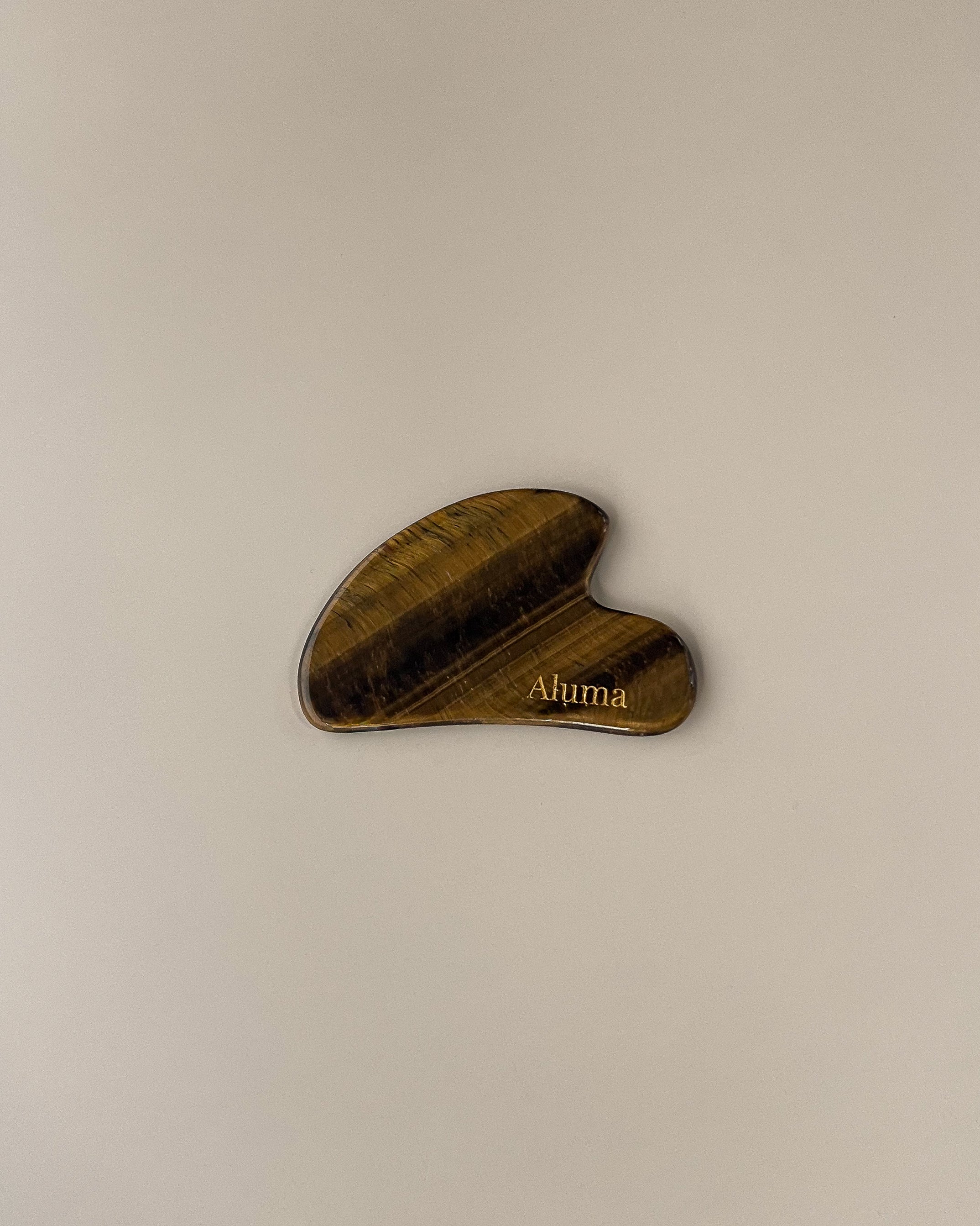 Yin gua sha i tiger eye sten fra Aluma på beige baggrund, varme gyldne årer der fremhæver stenens struktur til skulpturerende ansigtsmassage.