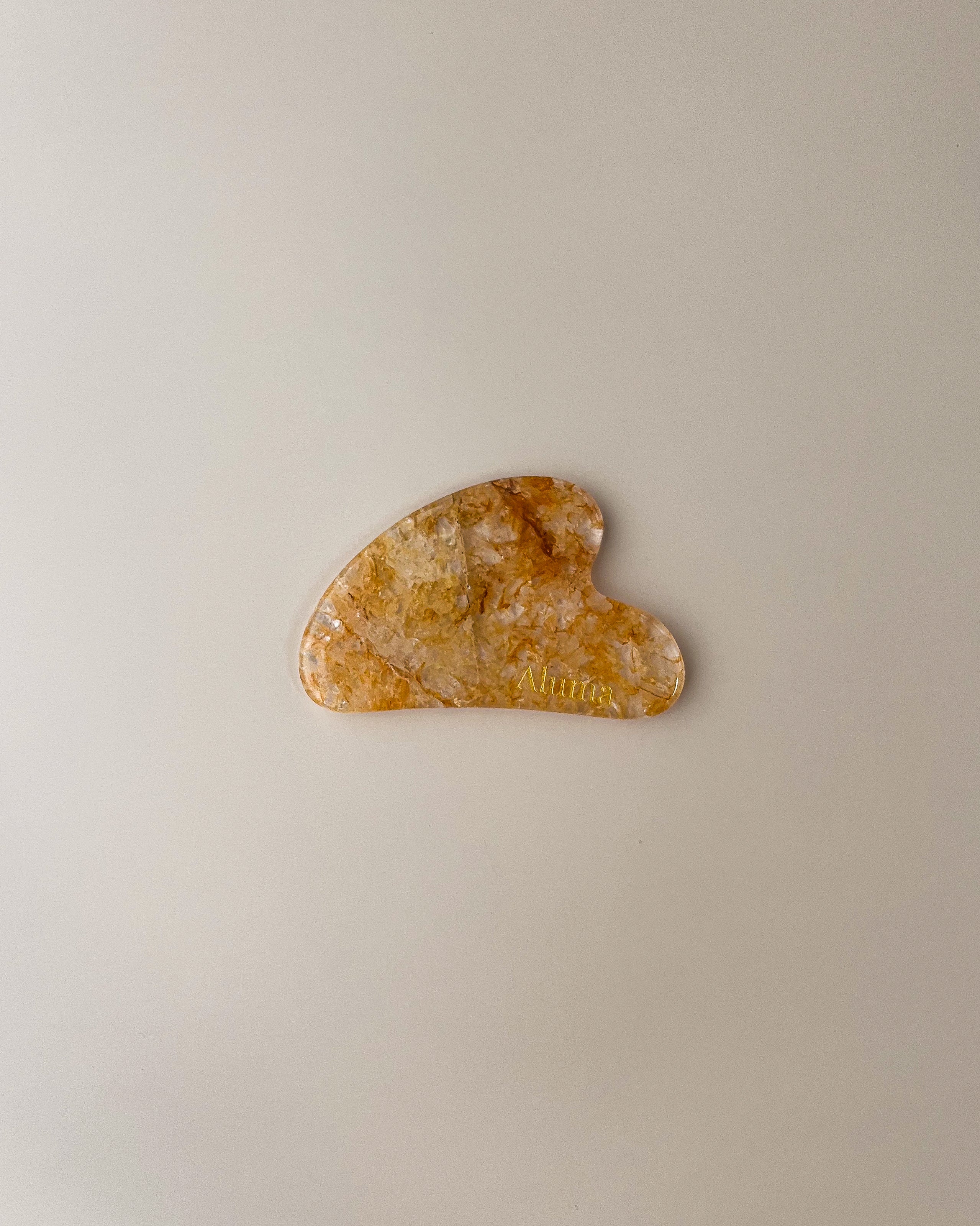 Yin gua sha i gylden yellow flower jade fra Aluma på beige baggrund, honningfarvet gua sha til ansigtsmassage med solvarm, glødgivende effekt.